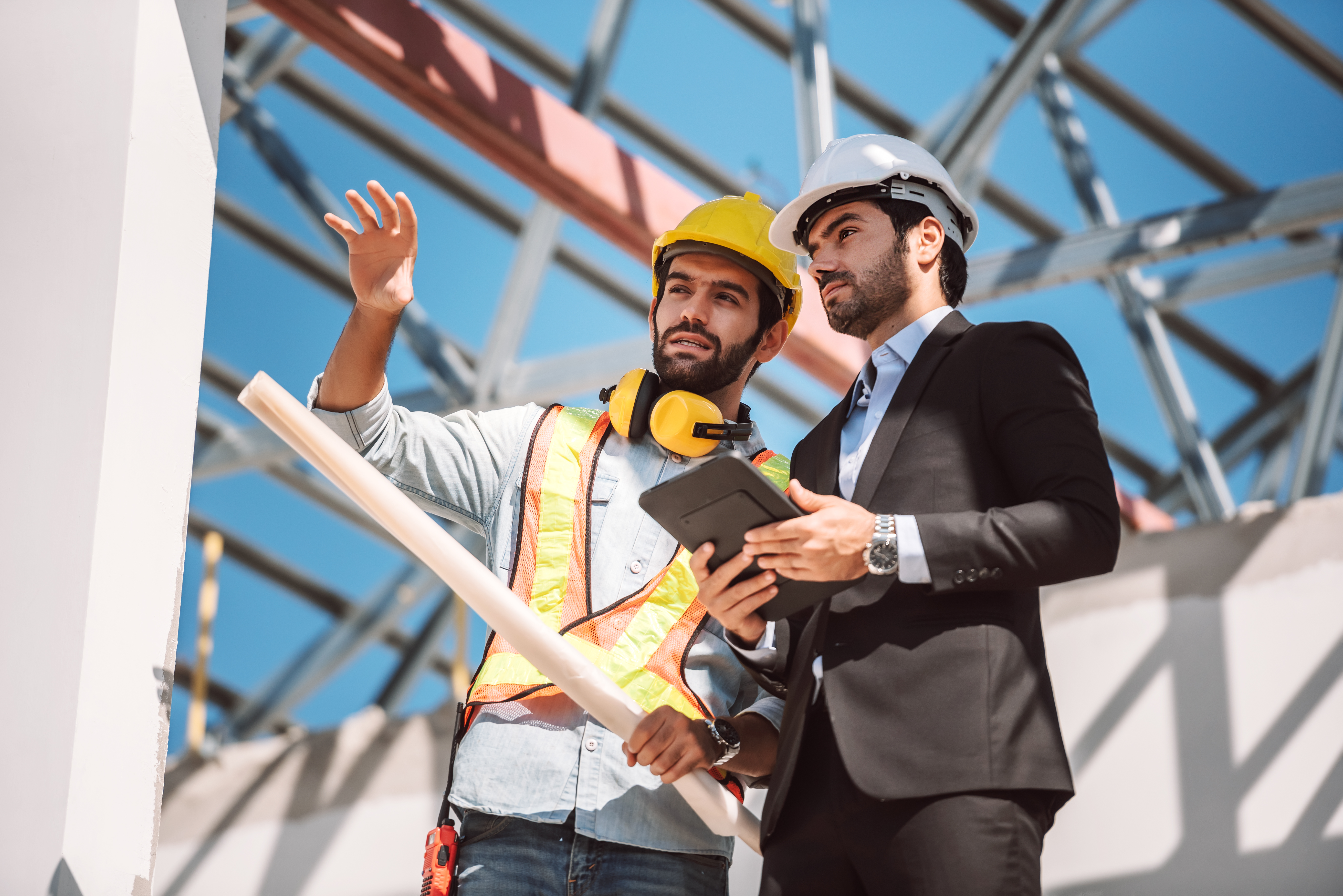 A construction worker wearing a white helmet, safety glasses, gloves, and a reflective vest with badges and tools stands outdoors at a construction site, helping to build a new home while looking off to the side.